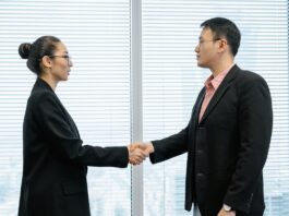 The head of a nonprofit wearing a long black coat shakes hands with a top donor in her office